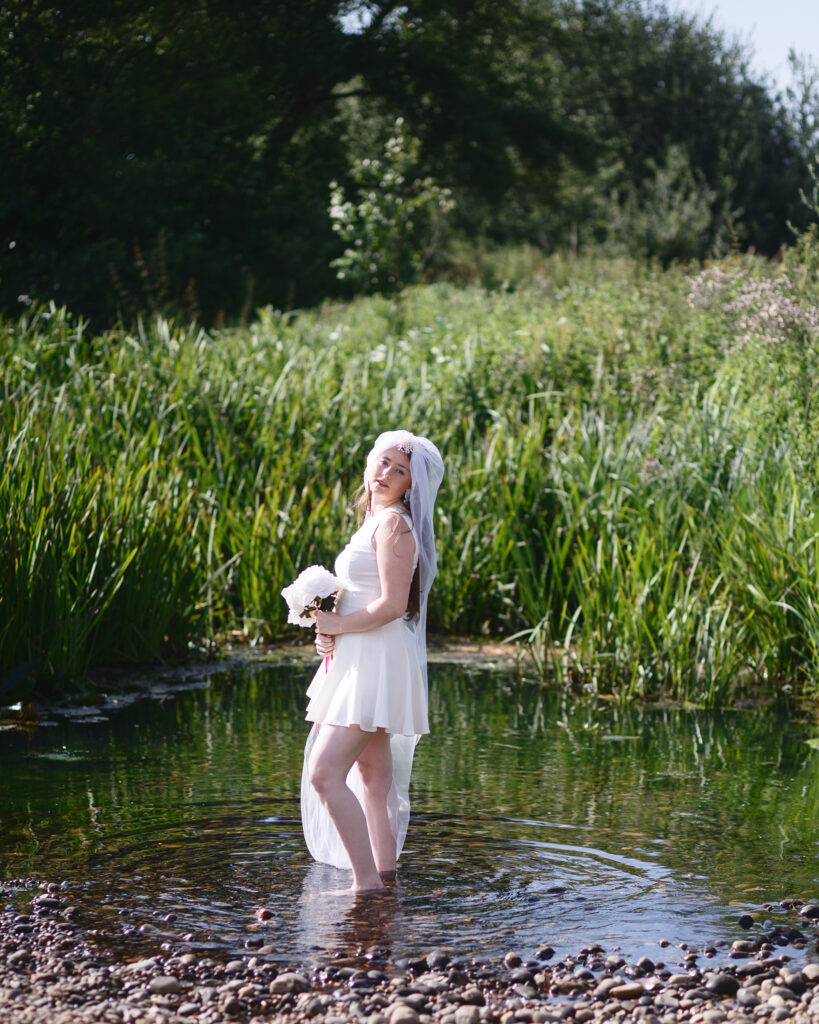 Sophia standing in the river at Halesworth in a cream dress with her veil trailing in the water