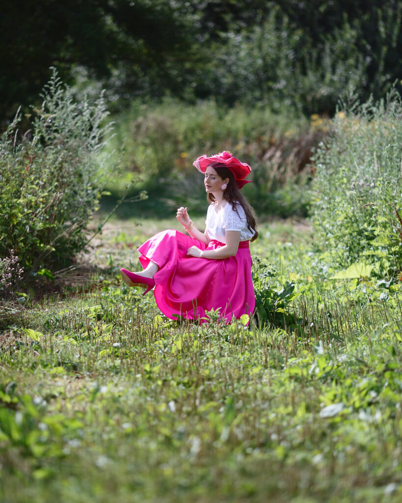 Sophia seated on a wooden bench in fuchsia pink with long pink gloves and a wide-brimmed hat